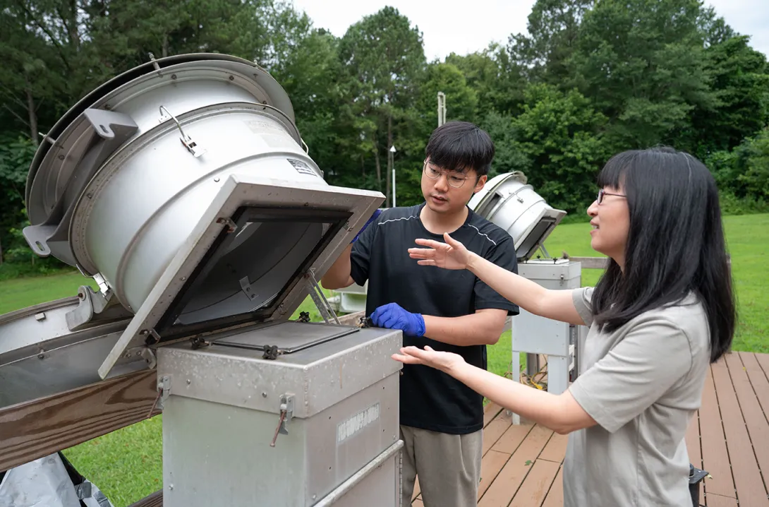 A male and female researcher working with a metal piece of equipment outdoors with trees and grass in the background