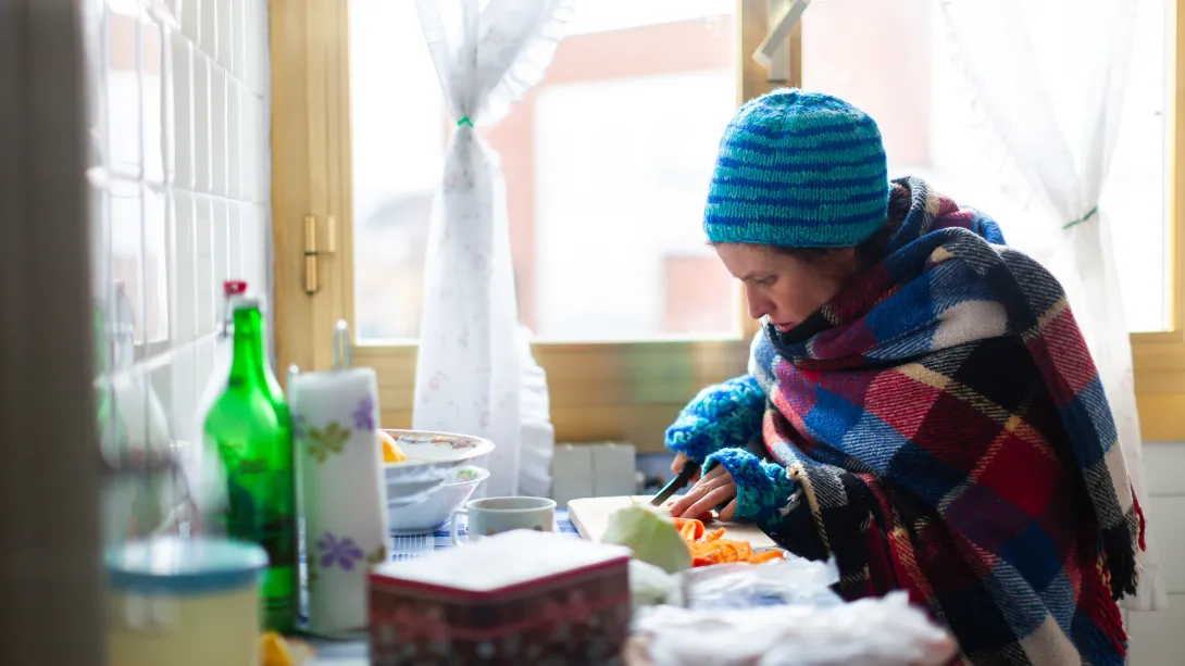A woman wearing a hat and warm clothing prepares food in her kitchen.