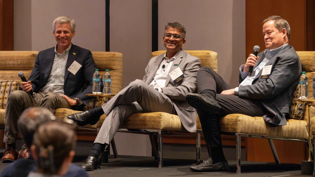 Three men sit on stage in a panel discussion, smiling and holding microphones. Water bottles rest on small tables beside their chairs.