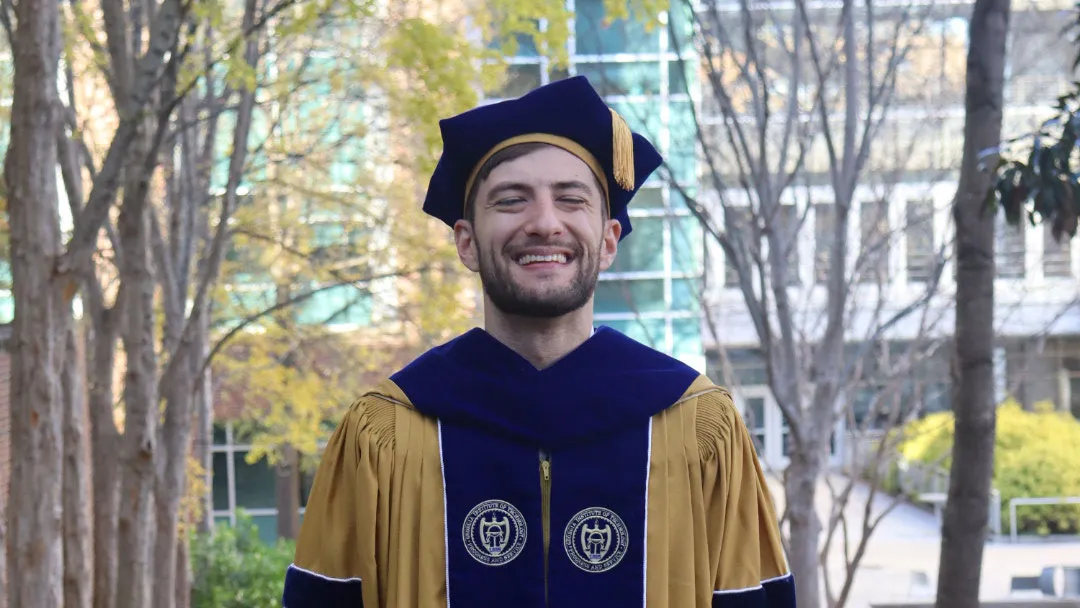 A smiling graduate stands on a tree-lined campus walkway covered with fallen leaves. He is wearing a gold doctoral gown with blue velvet panels and sleeve bars, along with a matching blue tam and tassel. Campus buildings and autumn trees are visible in the background.