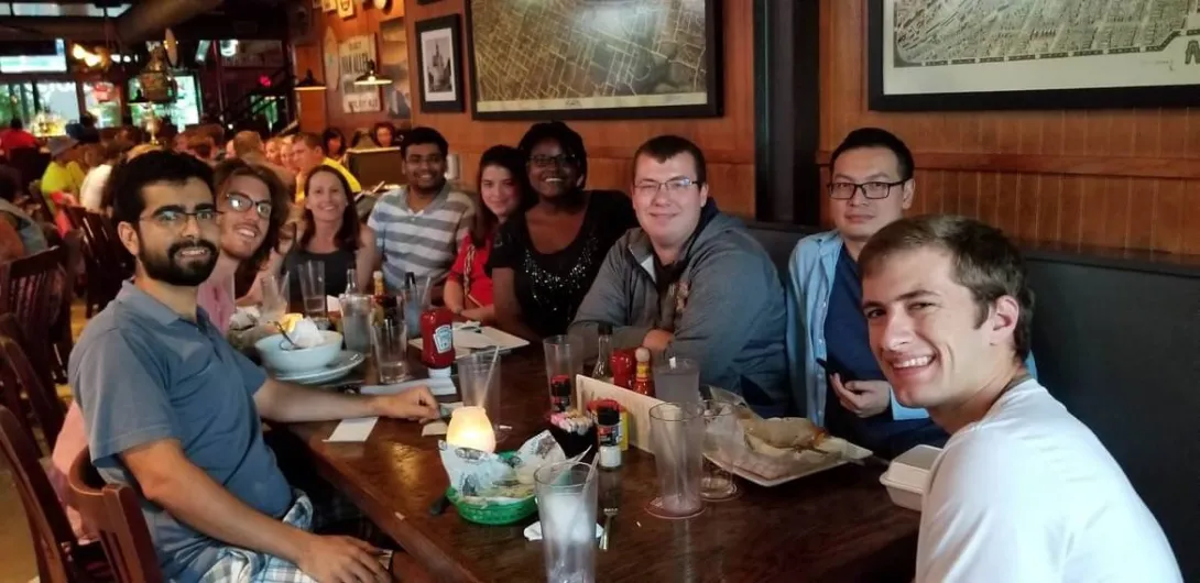 A group of nine people sit together around a long wooden table in a restaurant. Plates, drinks, and condiments are on the table. The group smiles toward the camera, with framed maps and warm lighting visible in the background.