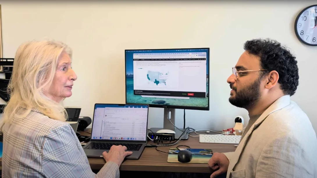 Two people talking at a standing desk with a monitor and laptop.