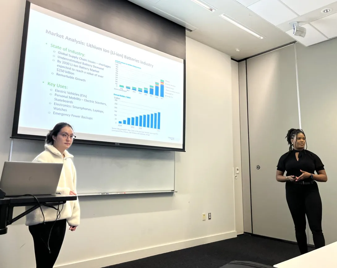 Two presenters in a classroom setting deliver a market analysis presentation, with slides showing charts and data projected on a screen.