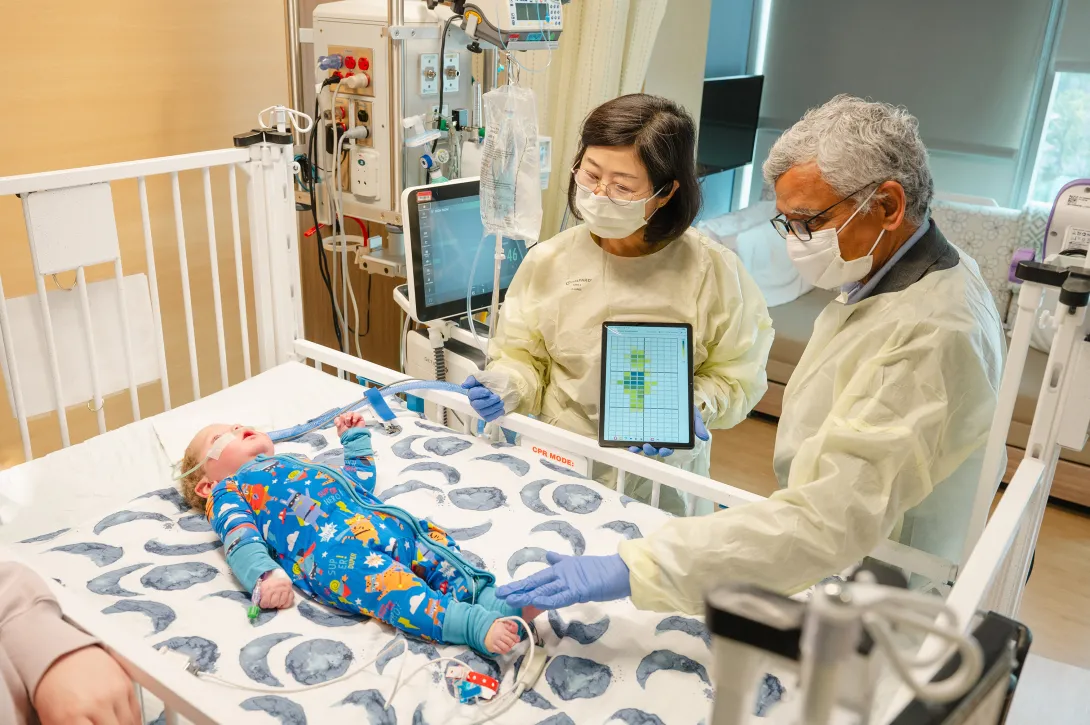 Two adults wearing protective gowns and gloves stand beside a hospital crib, using a tablet device while examining an infant lying on the mattress as medical equipment and monitors surround the crib.