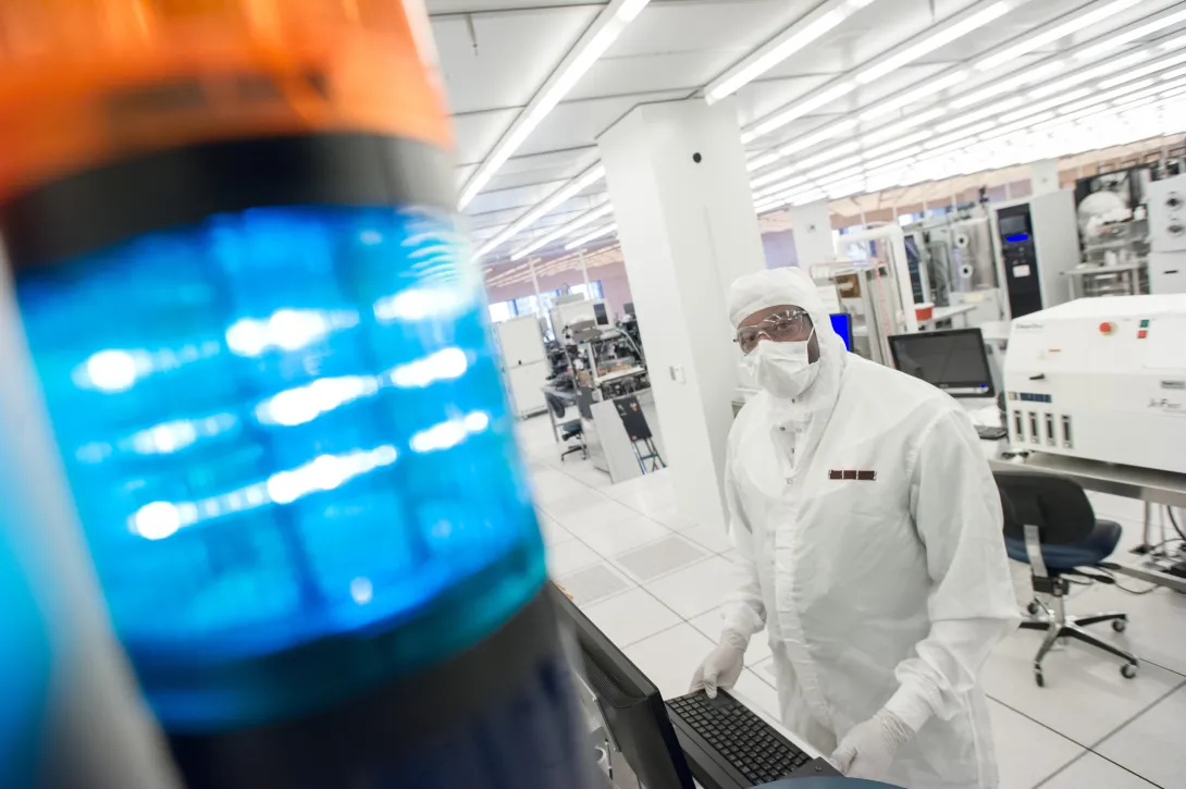 A cleanroom technician in protective gear works at a computer workstation in a semiconductor lab, with a blue signal light in the foreground and lab equipment behind them.