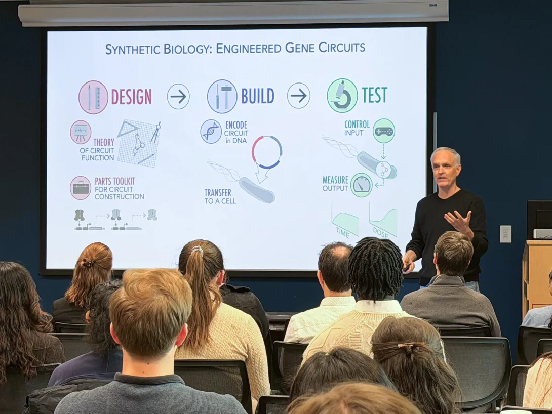 A presenter stands at the front of a lecture room speaking to a seated audience while a projected slide titled “Synthetic Biology: Engineered Gene Circuits” illustrates the design–build–test cycle with diagrams and icons explaining gene circuit construction and testing.