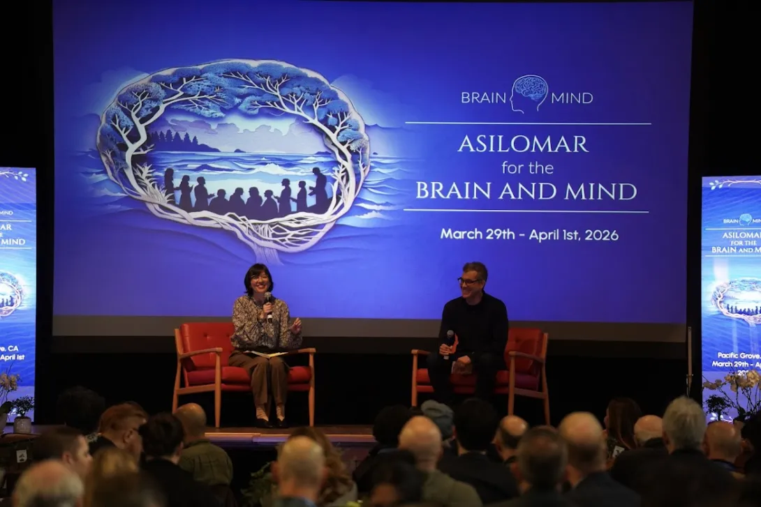 Seated on the left, Karen Rommelfanger speaks on a panel at the 2026 Asilomar for the Brain and Mind conference. Panelists sit on stage in front of a large screen displaying the conference name, dates, and a brain-themed graphic, with an audience visible in the foreground.