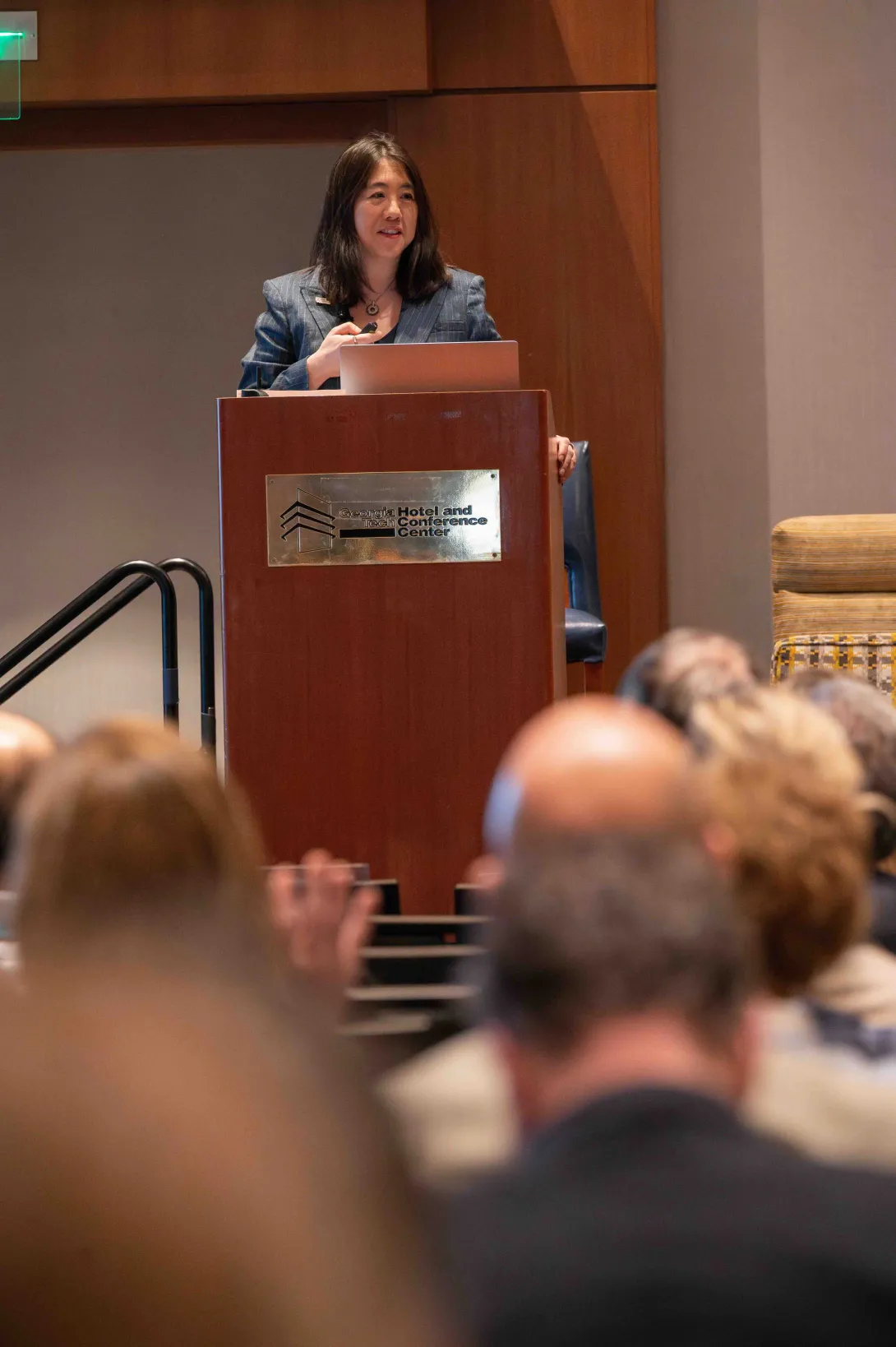 Vanessa Chan speaks at a podium at the Georgia Tech Hotel and Conference Center, addressing an audience. She holds a clicker and stands behind a laptop during a formal presentation.
