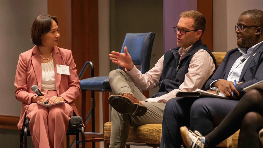Three panelists sit on stage during a discussion, with one man gesturing as he speaks while the others listen. The moderator holds a microphone and looks toward him.