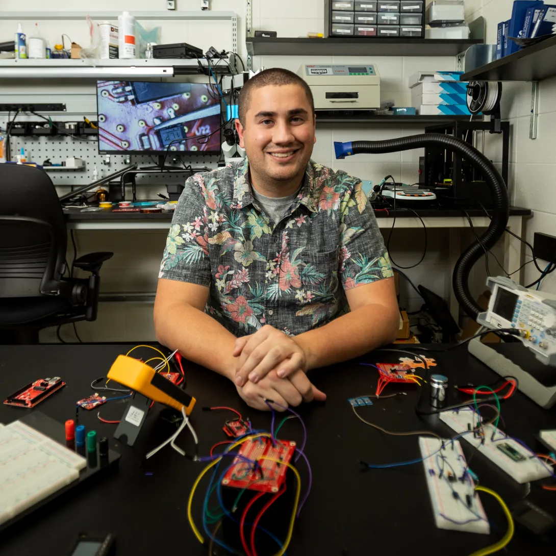 Josiah Hester sits at a desk in an electronics lab at Georgia Tech with an array of prototype projects and test equipment in front of him.