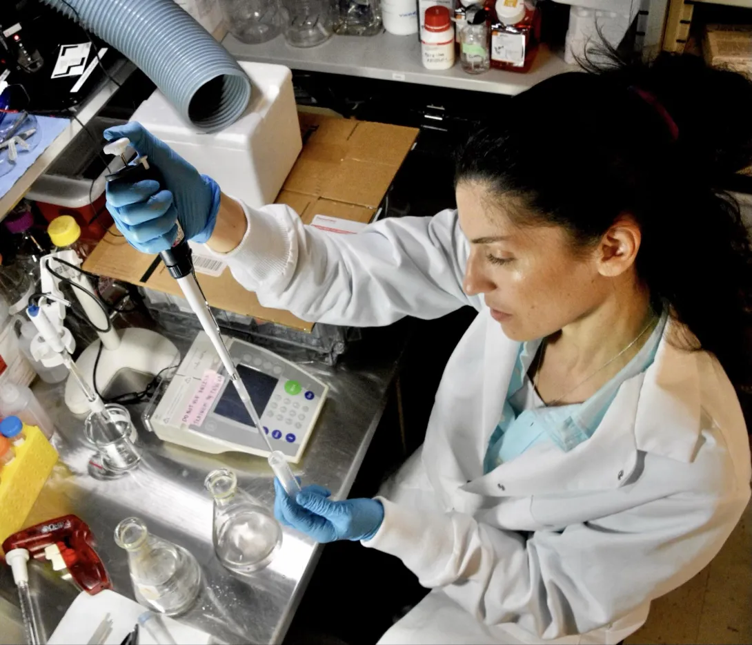 Picture of Patricia Stathatou wearing a white lab coat and blue latex gloves, holding a syringe and test tube