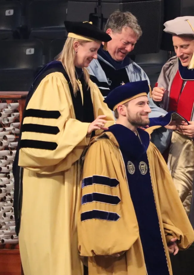 During a hooding ceremony indoors, a faculty member places a doctoral hood over the shoulders of a seated graduate wearing a gold gown and blue velvet doctoral regalia. Two additional faculty members in academic dress stand nearby, smiling. Rows of rolled diplomas are visible in the background.