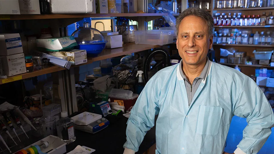 A man in a light blue lab coat standing at a laboratory bench with pipettes, containers, and scientific supplies on shelves behind him.