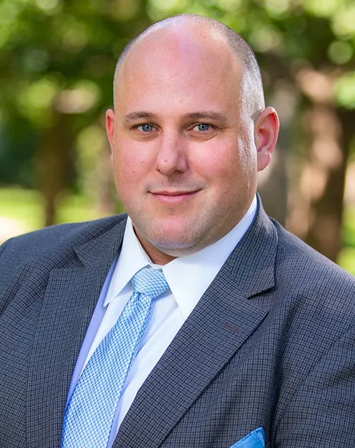 Head-and-shoulders portrait of a person wearing a dark suit jacket, light blue tie, and white shirt, photographed outdoors with trees and greenery softly blurred in the background.
