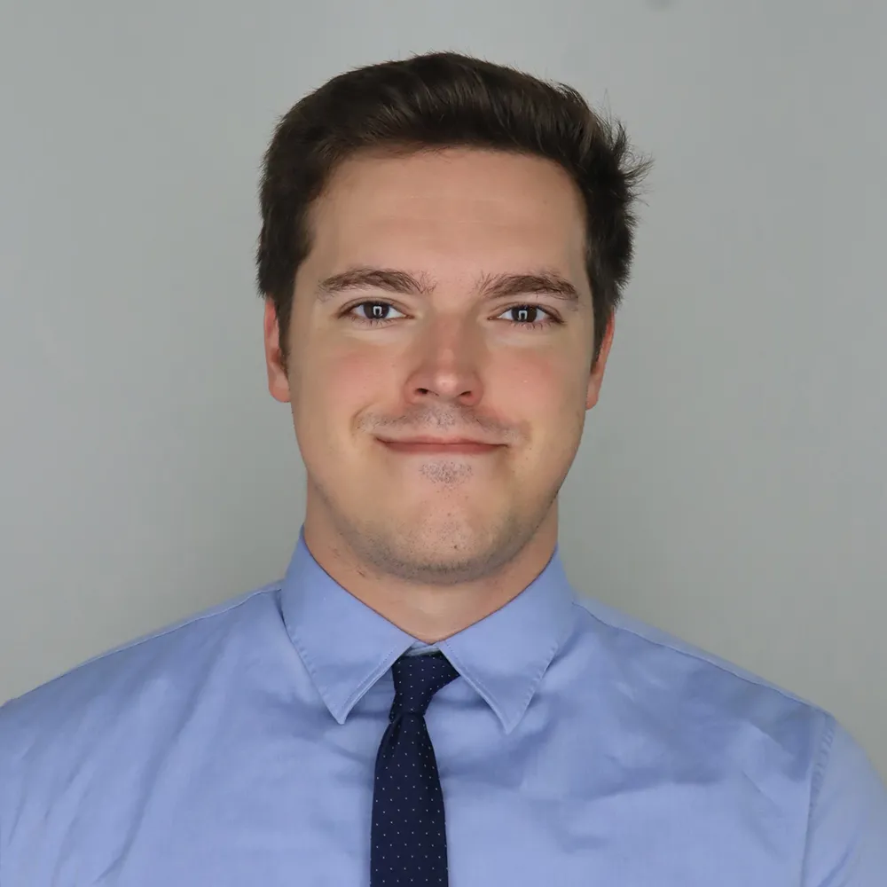 Headshot portrait of an individual photographed from the chest up against a plain light gray background. The individual is wearing a light blue button‑down shirt and a dark patterned tie, facing the camera in a centered studio-style composition. The image is evenly lit with a neutral backdrop and no visible text or additional objects.