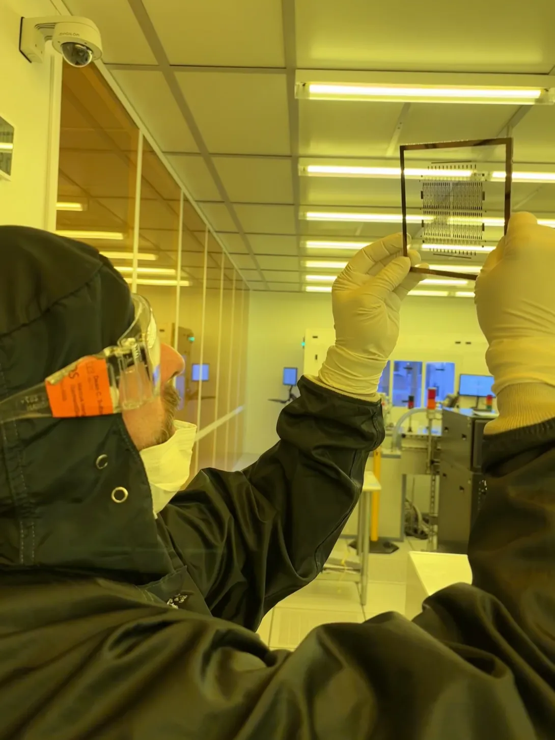Dean Sutter, wearing cleanroom protective gear and gloves, holds a semiconductor wafer up to the light inside a Georgia Tech research cleanroom.