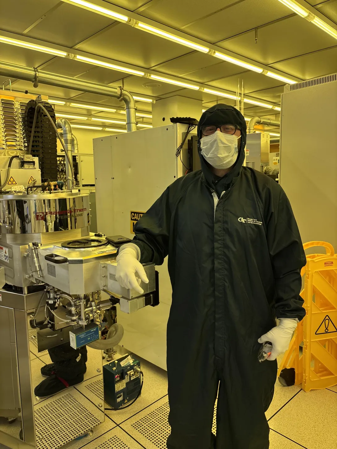 Dean Sutter stands beside semiconductor fabrication equipment in a Georgia Tech cleanroom while wearing full cleanroom protective attire and a face mask.
