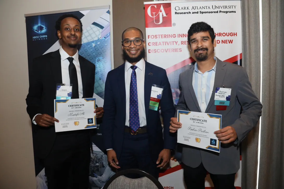 Three men in business attire stand side by side at a professional event, smiling at the camera. The men on the left and right hold certificates of award. Behind them are banners for the HBCU Chips Network and Clark Atlanta University’s Research and Sponsored Programs, highlighting innovation, creativity, and research.