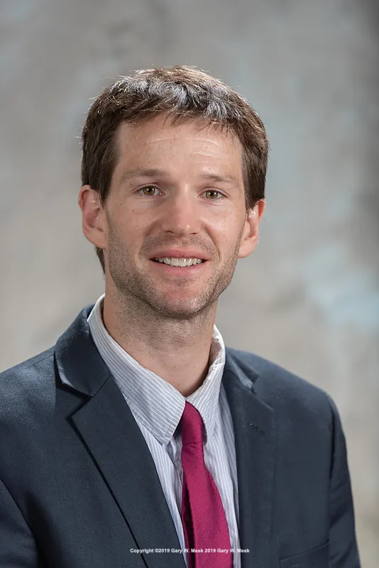 A man with brown hair and a short beard smiles for a portrait while wearing a dark blue suit and red tie.