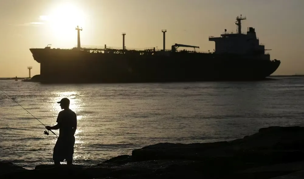 An oil tanker ship with the sun in the background and a man with a cap with a fishing poll in the foreground