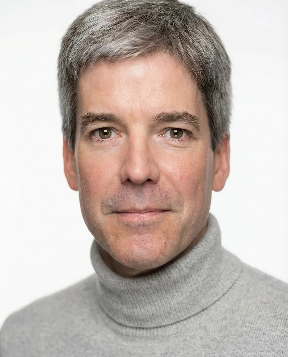 Headshot portrait of an individual photographed from the shoulders up against a plain light gray background, wearing a light gray turtleneck sweater. The individual faces forward with short, neatly styled hair and evenly lit features, centered in the frame.