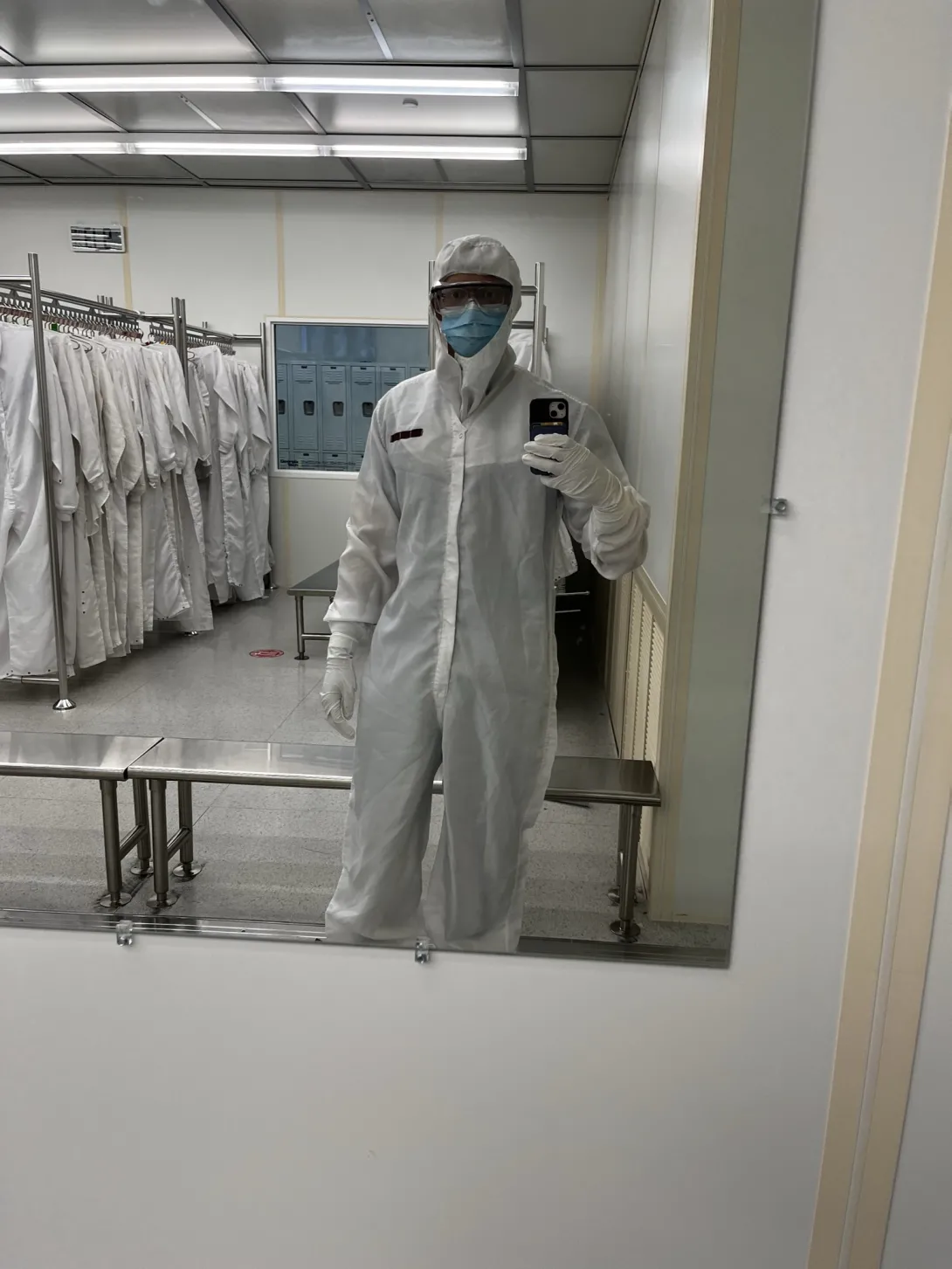 Calib Lanier wearing a full cleanroom suit, face mask, safety glasses, and gloves takes a mirror selfie in a gowning area lined with cleanroom garments and lockers.