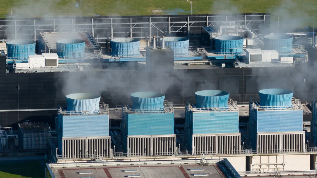 Aerial view of a large industrial facility with multiple blue cylindrical cooling towers arranged in rows, releasing visible steam into the air. The structures are connected by metal walkways, pipes, and equipment, with a darker building facade behind them. Green grass and patches of standing water are visible in the distance beyond the facility.