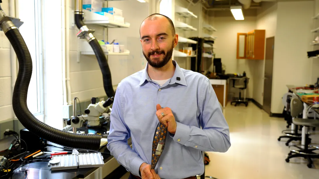 A researcher stands in a laboratory holding a flexible, transparent wearable device embedded with small electronic nodes, with microscopes and lab equipment visible in the background.