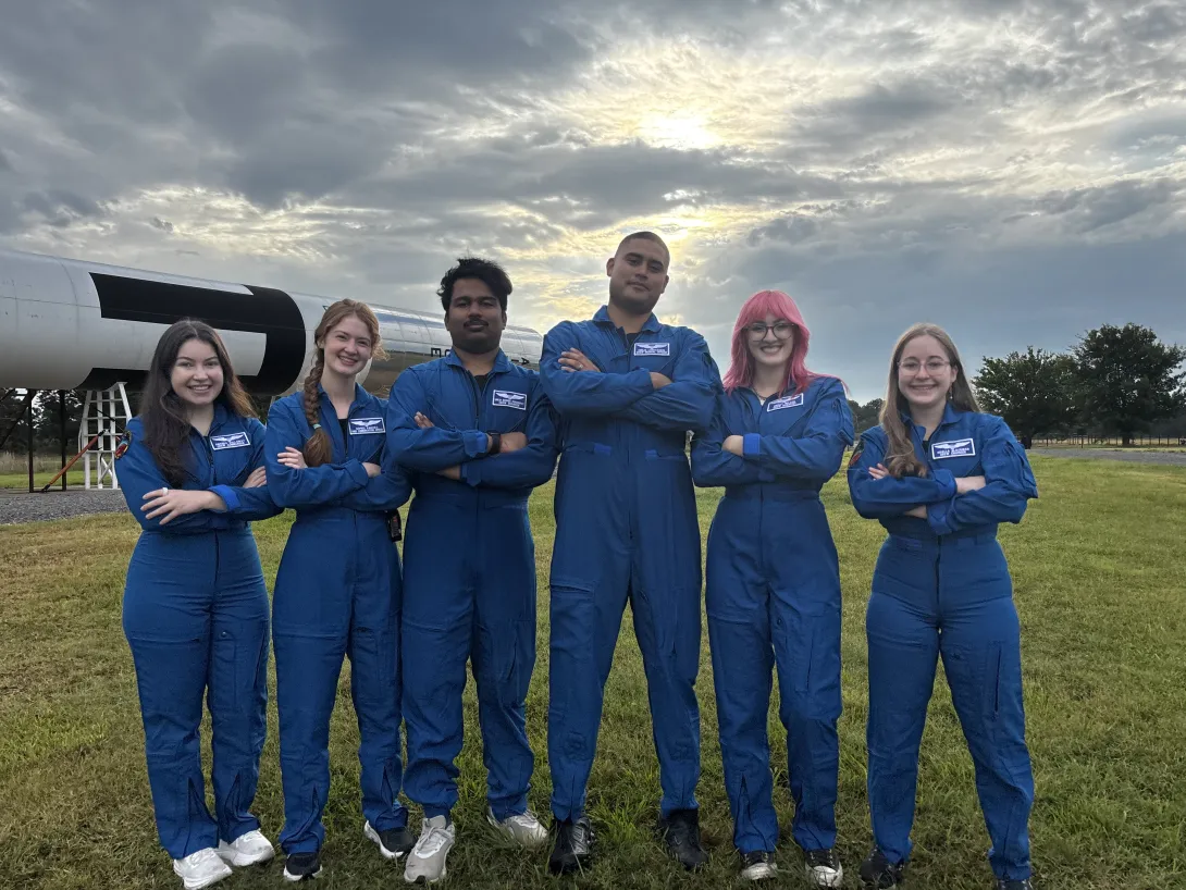 Five people wearing matching blue flight suits stand with arms crossed in a grassy field beside a large rocket display. Sunlight breaks through clouds overhead in the background.