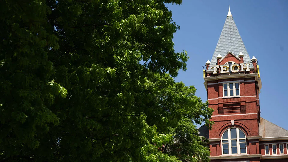 Georgia Tech’s historic Tech Tower rises above leafy green trees on a clear day, featuring a red brick facade, arched windows, and a pointed gray roof with the word “TECH” displayed prominently near the top.
