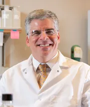 A man in a white lab coat and glasses, with a gold tie