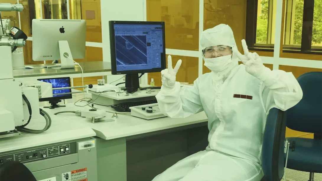 A researcher in a cleanroom suit, mask, and goggles sits beside a workstation with a microscope and computer, holding up peace signs.