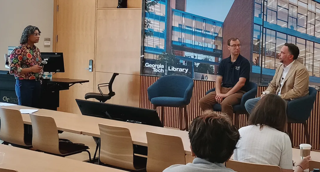 Panel discussion in a lecture room featuring two seated speakers at the front of the room and a presenter standing beside a lectern. A large display behind the speakers shows an image of the Georgia Tech Library building, while audience members are seated at tables facing the discussion. The room has light wood paneling throughout the space.