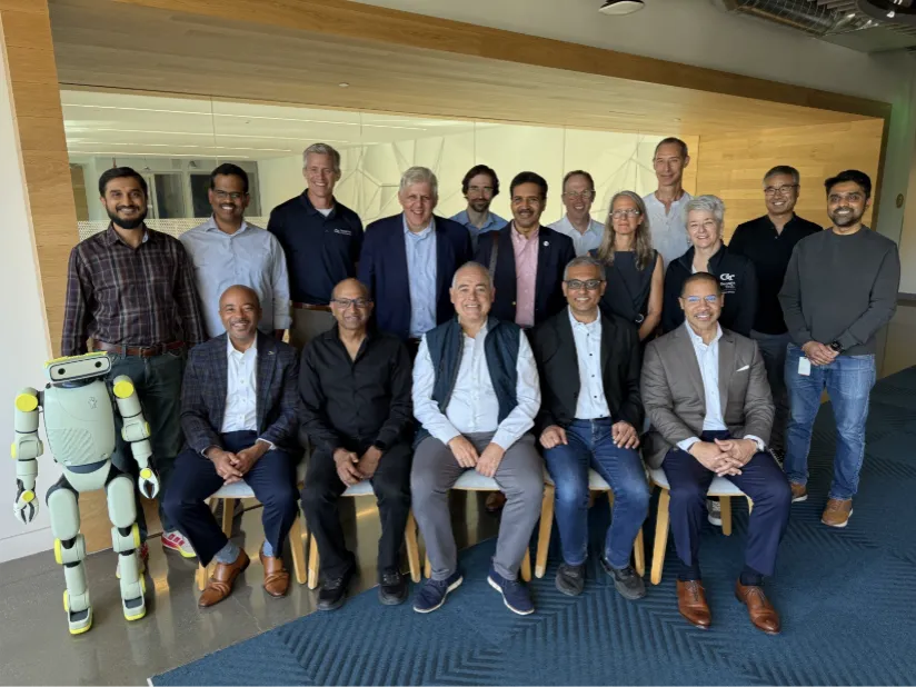Image shows a small humanoid robot standing beside 5 men who are seated, with GT President Cabrera in the middle, wearing a blue vest. Behind the seated group are 12 people standing, including 2 women.