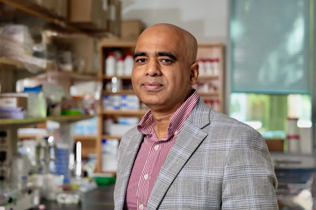 Ankur Singh, a man in a gray suit jacket with a dark pink button-up shirt stands in front of a work bench in a lab.