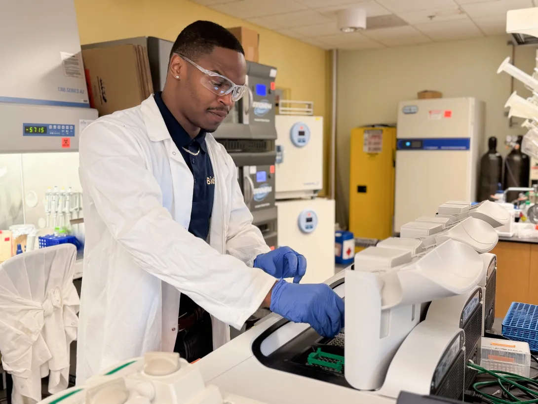A man in a lab coat wearing safety goggles and gloves puts samples into a machine in a scientific lab