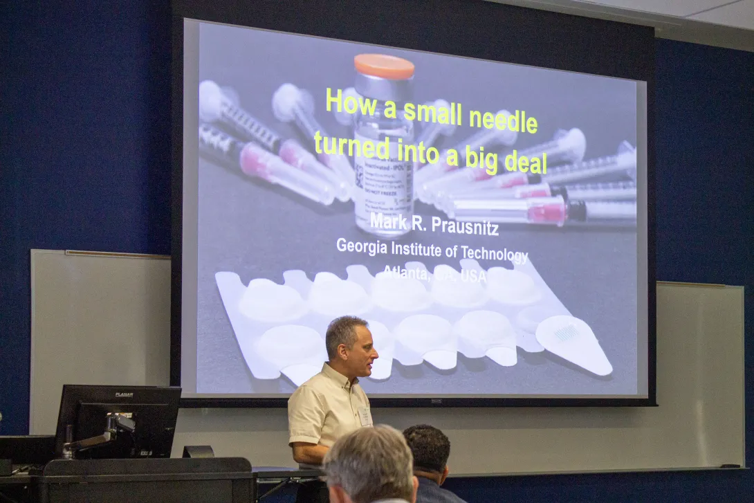 Mark Prausnitz speaks before a slide titled “How a small needle turned into a big deal,” featuring syringes, a vial, and microneedle patches.