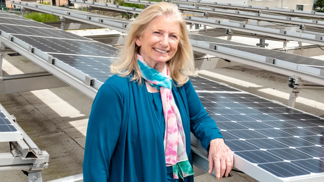 A woman with blonde hair and a blue sweater stands among solar panels. 