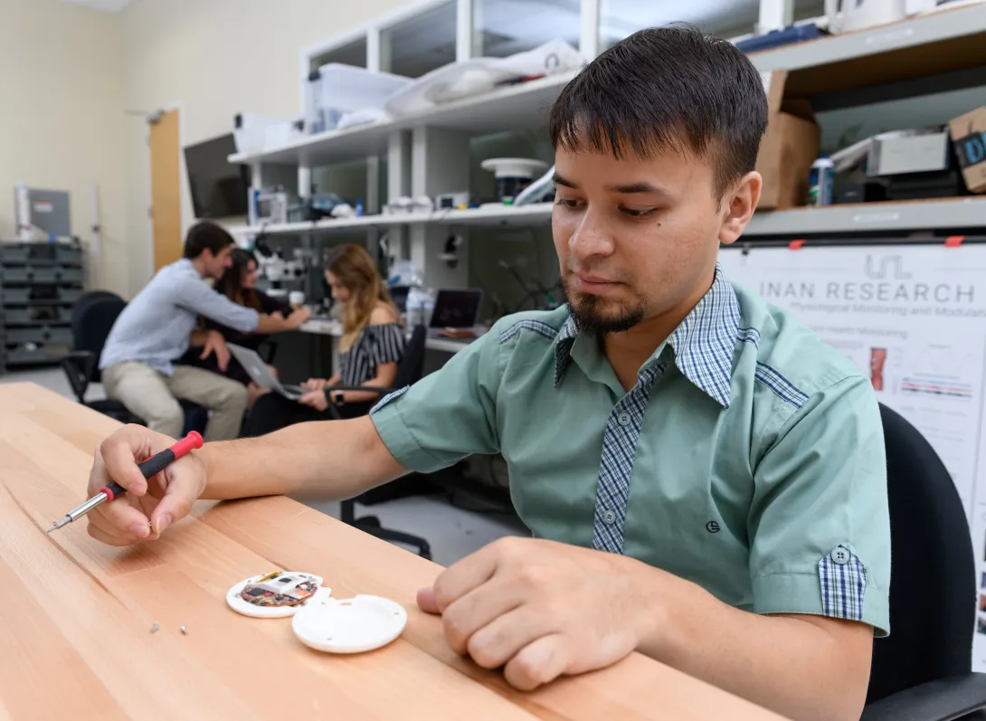 A man with tan skin and dark hair, wearing a mint-green shirt, is seated at a table and looking at the CardioTag device.