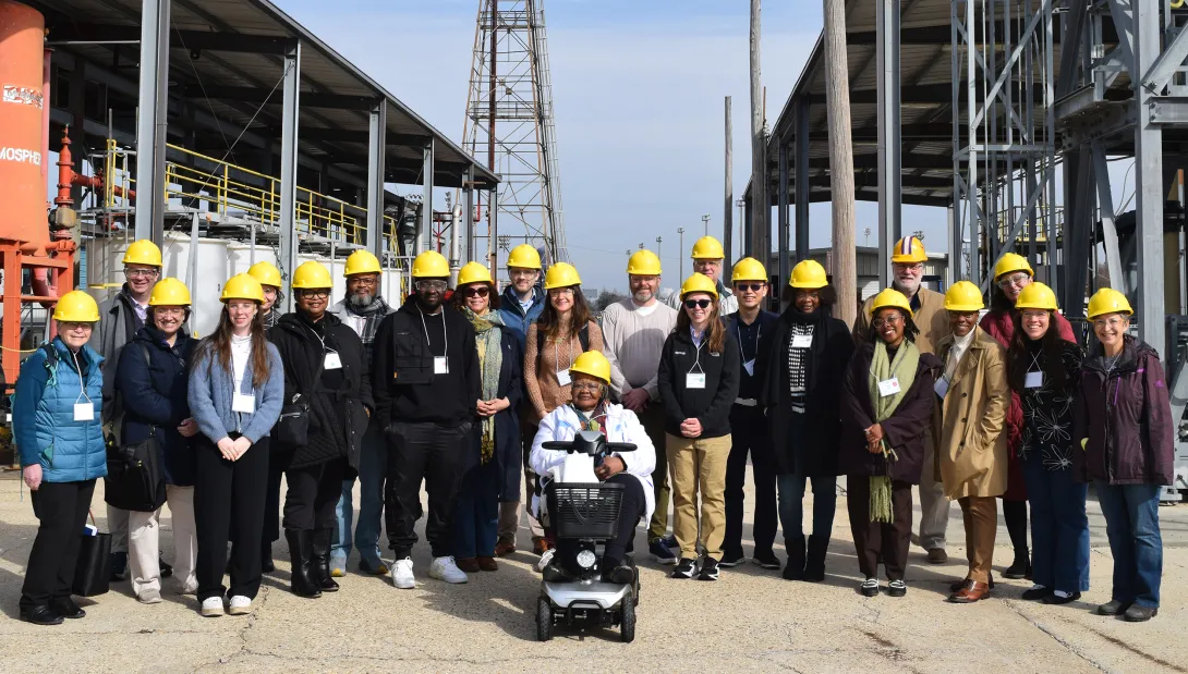 Group of about 20 people wearing yellow hard hats pose together at an outdoor industrial facility. One person sits on a mobility scooter in the front center, while the others stand behind and alongside, many wearing visitor badges. Metal structures, storage tanks, pipes, elevated walkways, utility poles, and a tall lattice tower fill the background.
