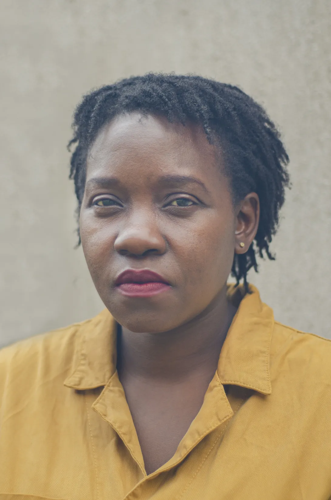 Photograph of a woman looking directly at the camera, she has short, dark hair in twists and is wearing a golden shirt with collar.