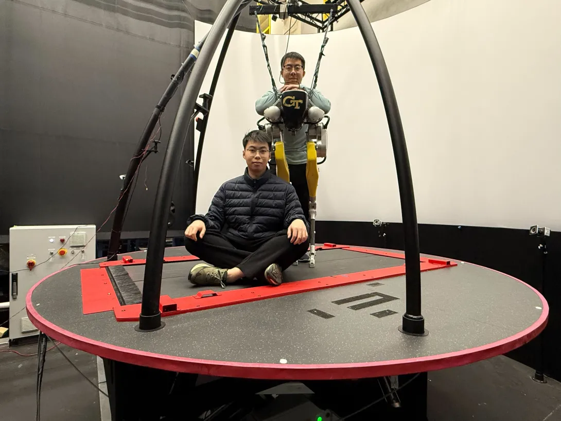 Two individuals on a large circular motion platform in a research lab, with one person seated cross‑legged on the platform and another suspended in a harness wearing a Georgia Tech exoskeleton system.